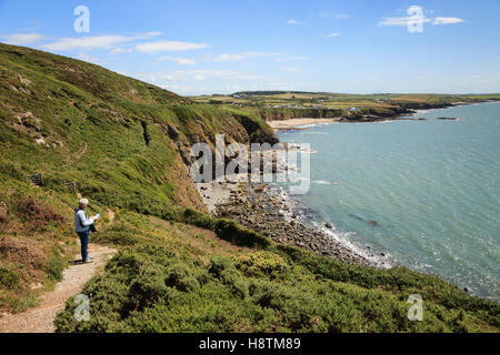 Walker with a map walking on coastal path with view along rugged coastline to Church Bay (Porth Swtan) Isle of Anglesey Wales UK Stock Photo