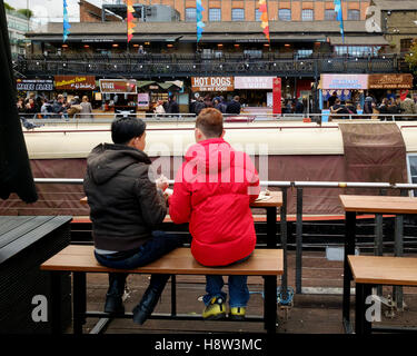 Two tourists sit on a bench in front of street food stalls at Camden Lock Market, Camden Town, London Stock Photo