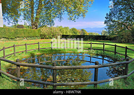 Pond circled by old wooden fence in a garden with mountains in the distant background. Stock Photo