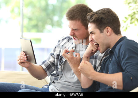 Two angry friends waiting for media content on line in a tablet with a slow connection sitting on a couch in the living room Stock Photo