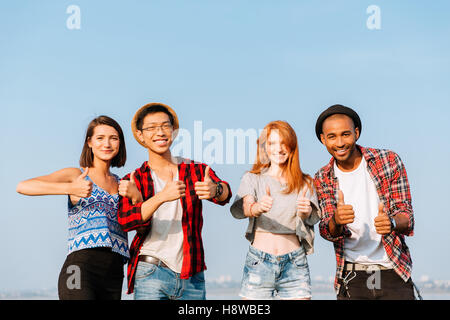 Multiethnic group of smiling young friends standing and showing thumbs up over blue sky background Stock Photo