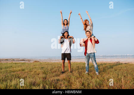 Two happy attractive young women sitting on their boyfriends shoulders with raised hands outdoors Stock Photo