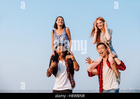 Two cheerful young women sitting on shoulders of their boyfriends outdoors Stock Photo