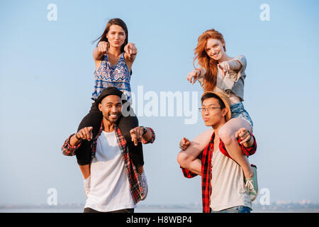 Two smiling young women sitting on shoulders of their boyfriends and pointing on you outdoors Stock Photo