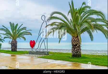 The sculpture of a man with the red heart in shopping cart, located on the seashore and surrounded by palms Stock Photo