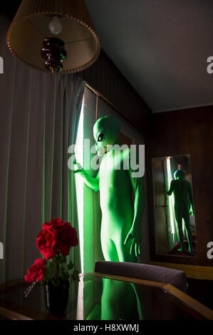 Green alien stands in a dark room pulling aside the curtains to look out the window Stock Photo