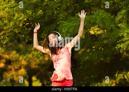 Happy teenage girl in red dancing in nature while listening to the music on headphones, smiling and looking at camera, on a sunny summer day. Stock Photo