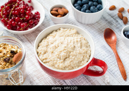 Porridge oats in red ceramic bowl. Served with red currants, fresh blueberries, almonds and muesli. White table background. Heal Stock Photo