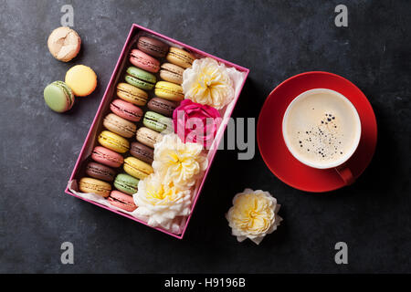 Colorful macaroons and coffee on stone table. Sweet macarons in gift box and flowers. Top view Stock Photo