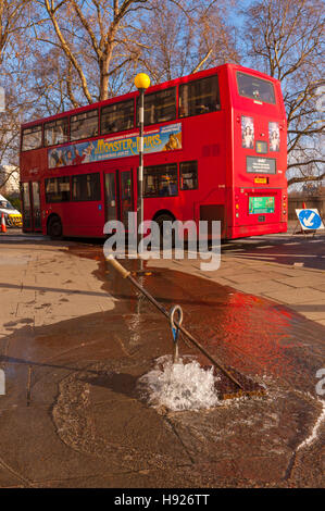 Red london bus driving past water flooding out of a manhole from a burst water main Stock Photo