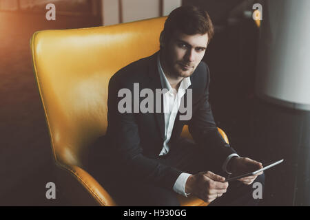Young serious successful man entrepreneur in formal business suite with a beard sitting on yellow armchair with digital tablet in office interior Stock Photo
