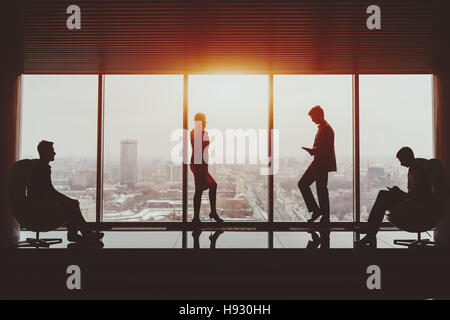 Silhouettes of businessman and businesswoman standing near window of skyscraper and two their male colleagues sitting on armchairs on opposite sides, Stock Photo