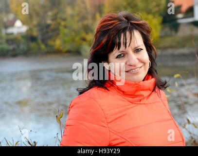 Portrait of smilling middle aged ordinary caucasian woman with long dark hair and red jacket in park. Stock Photo