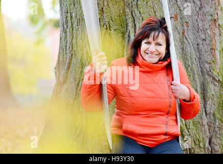 Portrait of smilling middle aged ordinary caucasian woman with long dark hair and red jacket sitting and swinging on swing in park. Stock Photo