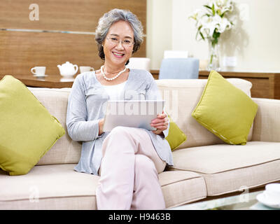 senior asian woman with tablet in hand sitting on couch at home looking at camera smiling Stock Photo