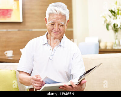 senior asian man sitting on couch at home reading a book, looking relaxed Stock Photo