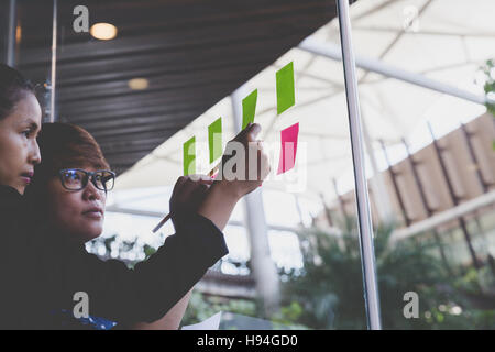 young businesswoman with colleague sticking adhesive notes on window in office Stock Photo