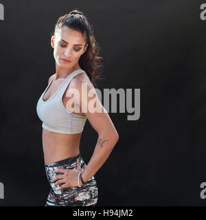 Slim and fit young female model in sportswear standing with her hands on hips. Muscular young woman standing on black background. Stock Photo