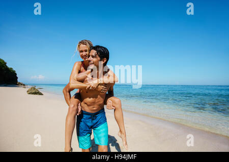 Portrait of happy young couple piggyback on tropical beach. Man carrying girlfriend on his back along the sea shore. Stock Photo