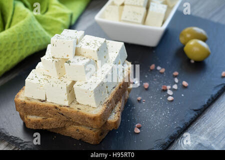 Tofu feta cheese with fresh oregano on a whole grain toast with linseed along with himalayan salt and two green olives on a black stone tray. Stock Photo