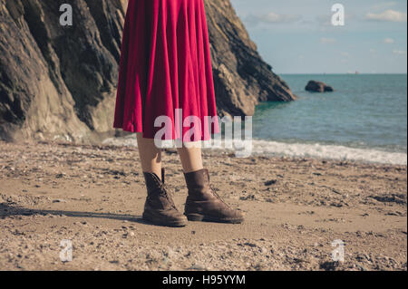 A young woman wearing a red dress and hiking boots is standing on the beach Stock Photo