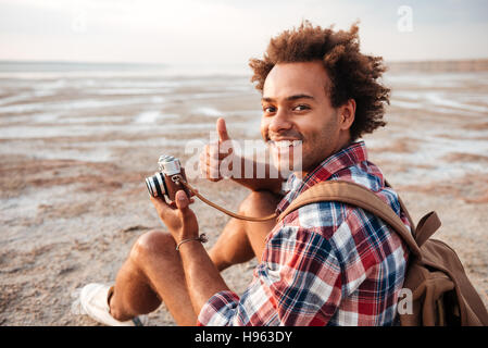 Happy african american young man with backpack taking photos and showing thumbs up on the beach Stock Photo