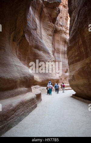 Tourists walking through the Siq, past ancient wall carvings, towards the entrance to Petra, Jordan Stock Photo