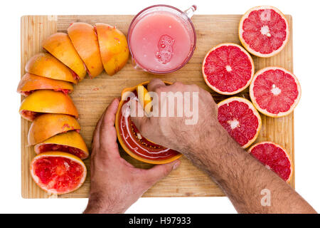Hands of a man squeezing fresh ruby grapefruit halves on a manual juicer with fresh cut juicy fruit on one side and squeezed skins on the other on a b Stock Photo