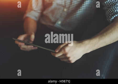 View of hands of young man in checkered shirt sitting on black leather sofa and working on digital tablet Stock Photo