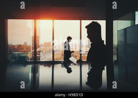 Silhouette of two businessmen working with their gadgets in office interior of skyscraper, man in front with digital tablet and man behind with phone Stock Photo