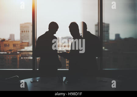 Silhouettes of two businessman near window and table of dark office interior, discussing something and looking on screen of digital tablet, cityscape Stock Photo