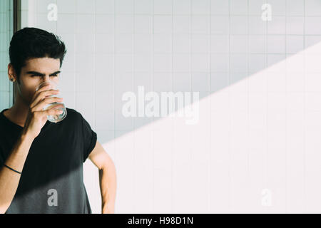 Handsome brazilian guy with earring, drinking cold water from transparent glass on the balcony near window, ceramic tiles behind with diagonal shadow Stock Photo