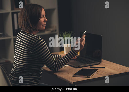 Business woman working overtime in the office, adult caucasian female sending text message from her smartphone while completing job task on laptop com Stock Photo