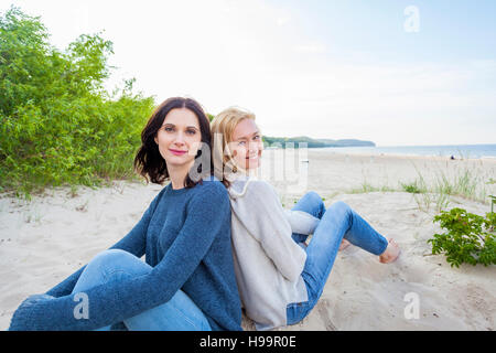 Two women on sandy beach sitting back to back Stock Photo