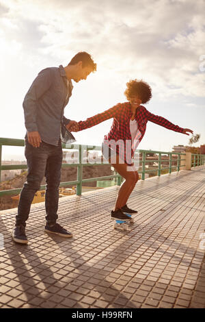 Curly haired girl learning to skateboard while holding boy holds her hand tightly in great concentration while she looks down with her arms out Stock Photo