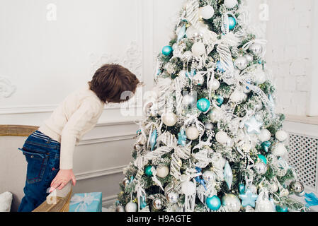 Funny little boy standing on a sofa near Christmas tree with colorful lights and looking to his reflection at xmas ball Stock Photo