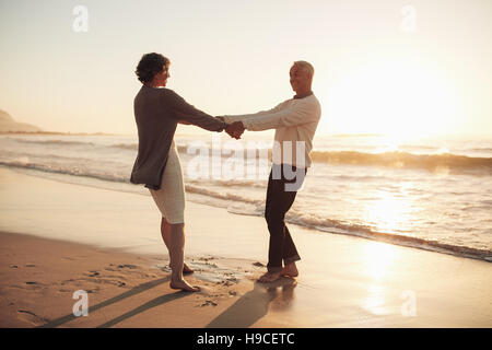 Full length outdoor shot of senior couple holding hands standing on the beach. Mature man and woman playing on the seashore at sunset. Stock Photo