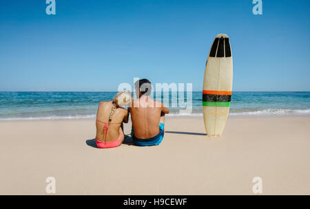 Rear view shot of young man and woman sitting on the beach with surf board. Couple relaxing on the sea shore on a summer day. Stock Photo