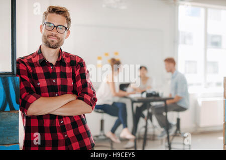 Portrait of relaxed young man standing in office with colleagues meeting in background. Stock Photo