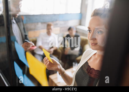 Creative business woman sticking adhesive notes on glass wall in office. Female in meeting with corporate colleagues. Stock Photo