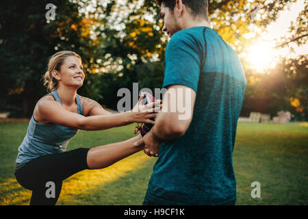 Male physical trainer assisting woman with leg exercise. Young woman being assisted by personal trainer in stretching exercises at park. Stock Photo