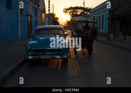 A horse and cart passes a classic US vintage car pared on a street in Cienfuegos Cuba with the sun setting behind Stock Photo