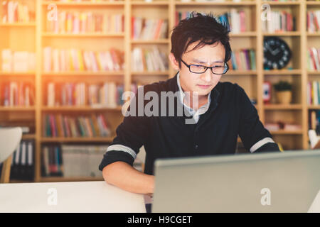 Young pensive Asian man working on laptop at home office or library with serious face, bookshelf blur background with copy space Stock Photo