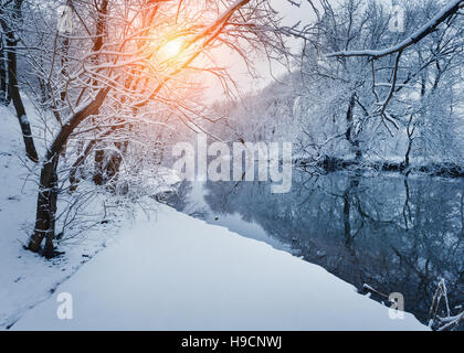 Winter forest on the river at sunset. Colorful landscape with snowy trees, frozen river with reflection in water Stock Photo