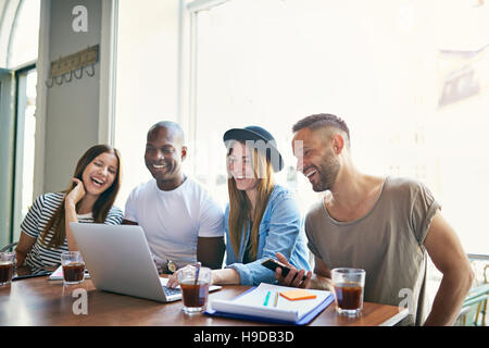 Four young smiling co-workers sitting at desktop and looking at laptop in light room. Stock Photo