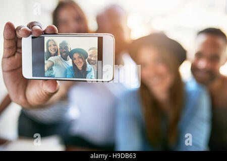 Close up phone in male hand making self portrait of four smiling people sitting in light room. Stock Photo