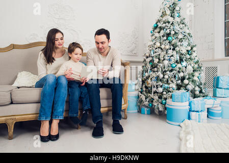 Cheerful family sitting in the living room having fun with the digital tablet that Santa Claus brought her, behind the decorated christmas tree Stock Photo
