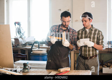 Happy male carpenter showing something to coworker at his notebook papers in workshop Stock Photo