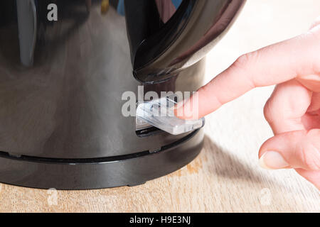 Close up of a manicured finger pressing the clear plastic switch of a black kettle on a wooden surface Stock Photo