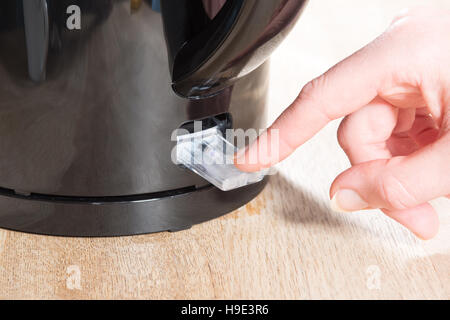 Close up of a manicured finger pressing the clear plastic ON button of a black kettle on a wooden surface Stock Photo